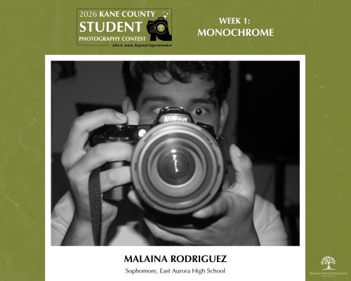 Monochrome portrait of a photographer aiming a Nikon DSLR camera straight at the viewer.