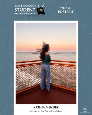 Person facing away from the camera, hair blowing and standing on a ferry's corner deck as the sun sets behind a cityscape across the water.